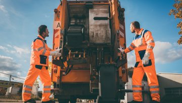 Two refuse collection workers loading garbage into waste truck