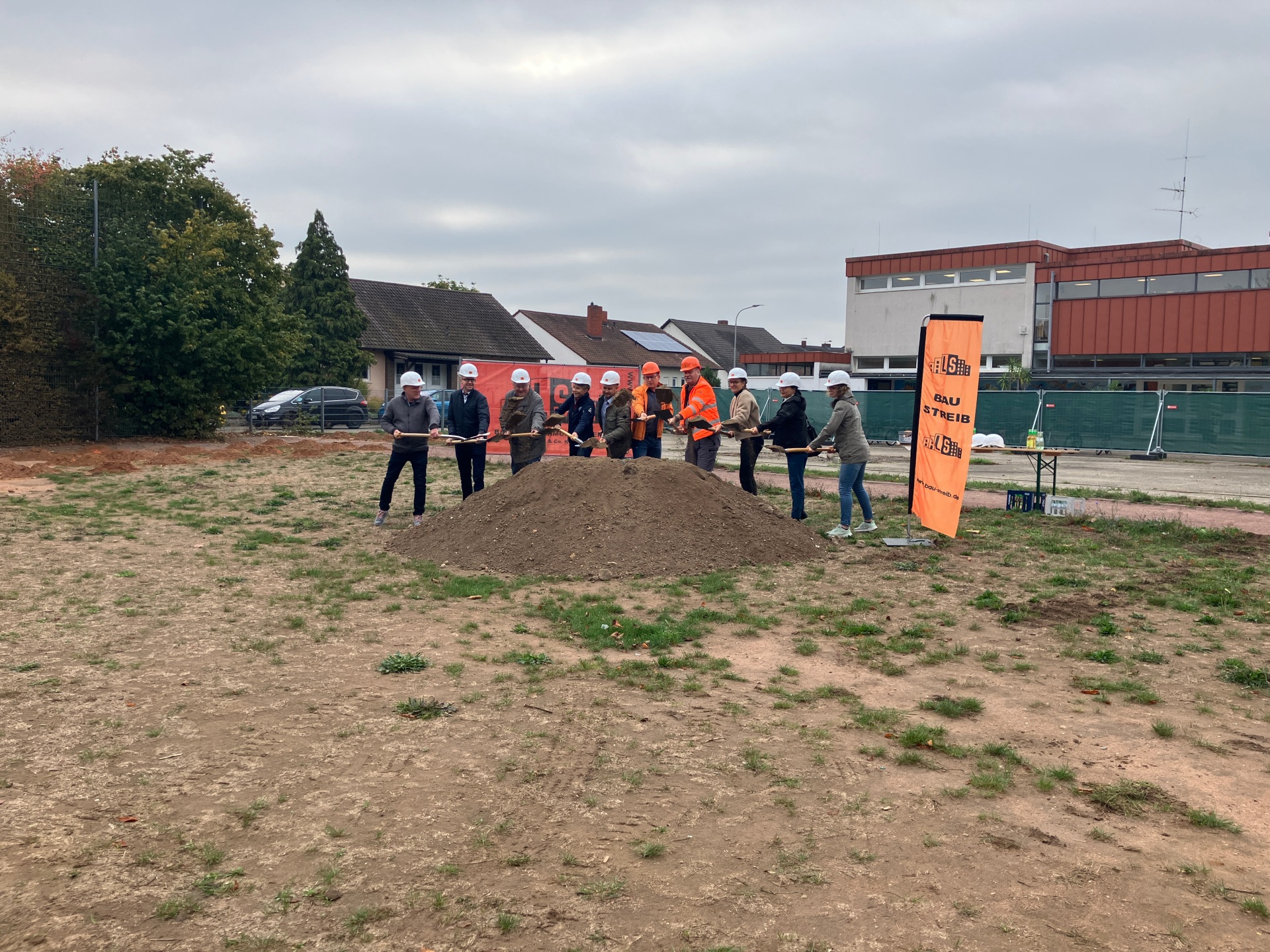 Gemeinsamer Spatenstich Eine Gruppe von Personen mit Bauhelmen und teilweise orangefarbener Warnkleidung steht gemeinsam mit Spaten an einem Erdhügel auf dem Gelände des geplanten Mensaneubaus. Im Hintergrund sind Bäume, Wohnhäuser und Schulgebäude zu sehen.